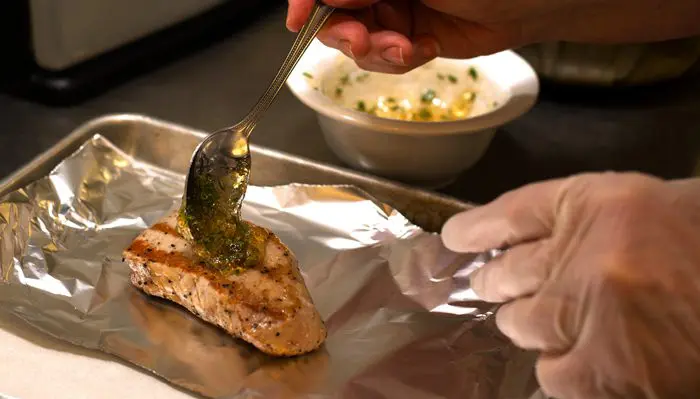 Hands in gloves apply seasoning to a piece of cooked fish on a baking tray.