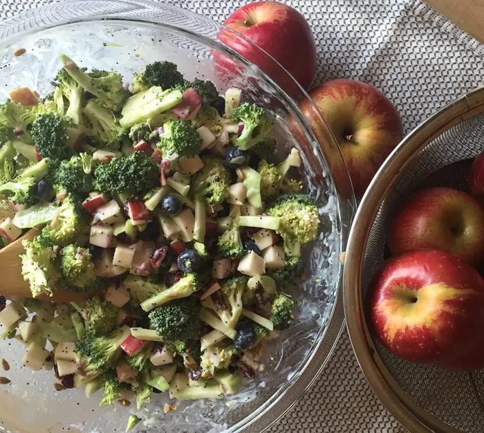 A bowl of broccoli salad beside fresh red apples on a woven placemat.