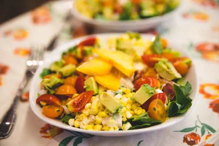 A colorful plate of salad with corn, tomatoes, mango, avocado, and greens on a floral tablecloth.
