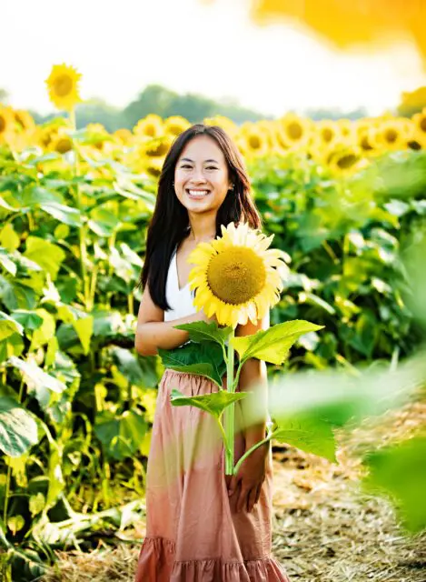 A smiling person holding a sunflower in a vibrant sunflower field.