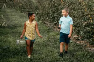 Two children are walking and smiling through a lush orchard with one holding a small sign.