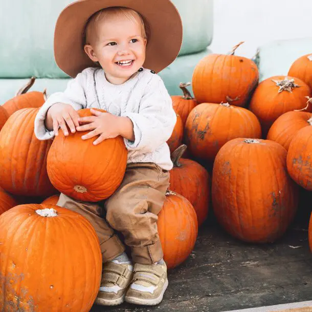 Smiling Child Holding Pumpkin with pumpkin display wearing cowbow hat.