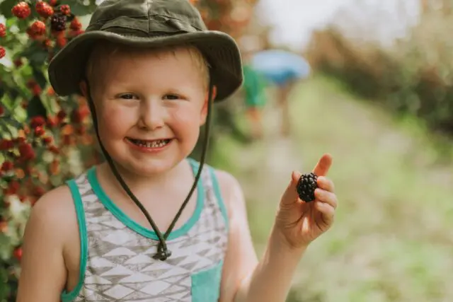 A smiling child wearing a bucket hat shows off a berry with someone in the background amidst greenery.