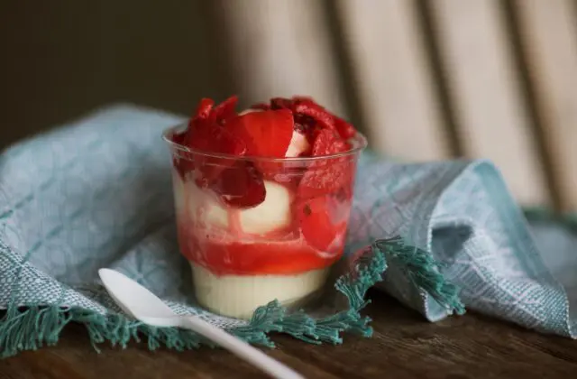 A cup of layered dessert with a creamy base and red topping sits next to a white spoon on a teal napkin.