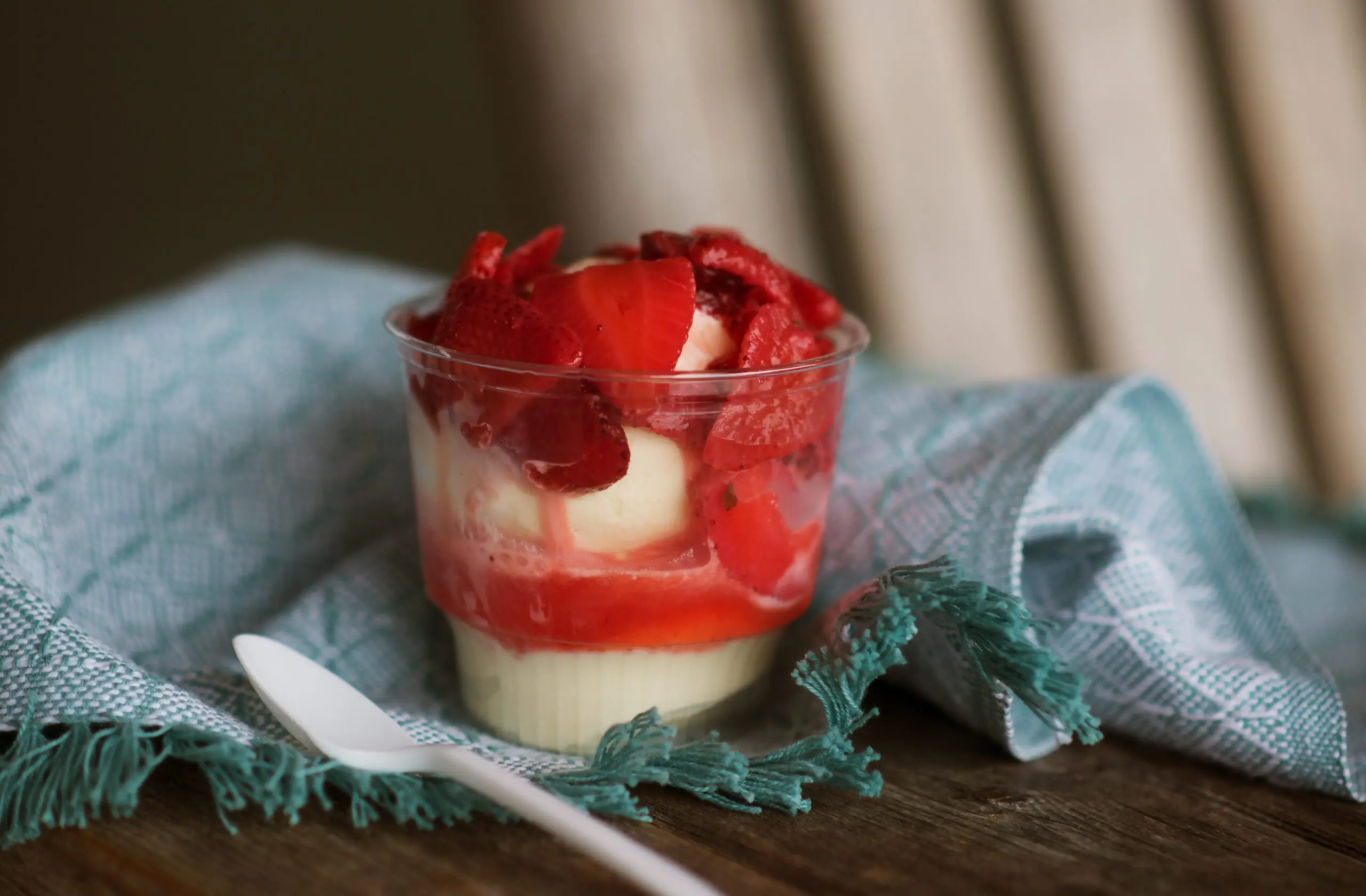 A cup of layered dessert with a creamy base and red topping sits next to a white spoon on a teal napkin.