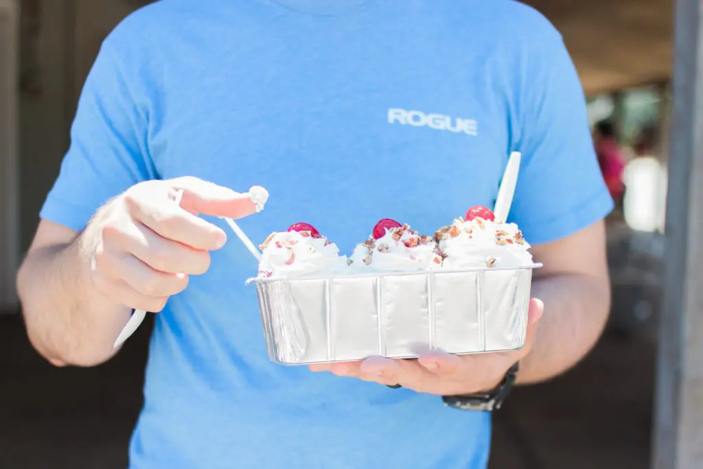 A person in a blue shirt is holding a tray with six scoops of ice cream topped with whipped cream and a cherry.