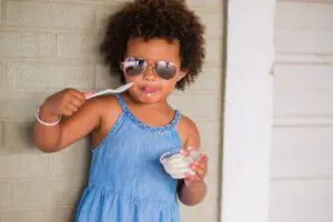 A young child in a blue dress and sunglasses enjoys yogurt with a spoon outdoors.