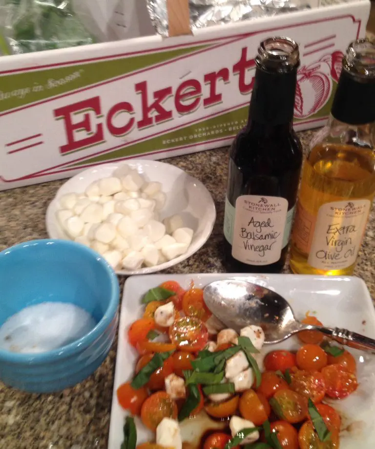 A variety of items are arranged on a countertop, including a bowl of cherry tomato salad, two bottles—one labeled "Apple Balsamic Vinegar" and the other "Extra Virgin Olive Oil"—a bowl of what appears to be sugar or salt, and a small bowl of small white objects, possibly mini mozzarella balls or marshmallows, all with a sign reading "Eckert's" in the background.