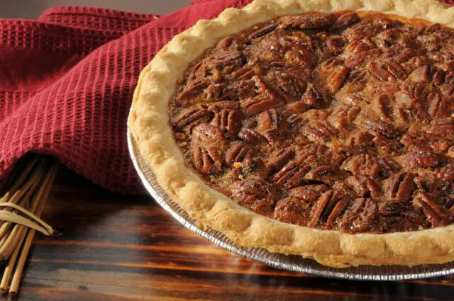 A freshly baked pecan pie sits on a wooden table next to cinnamon sticks and a red cloth.