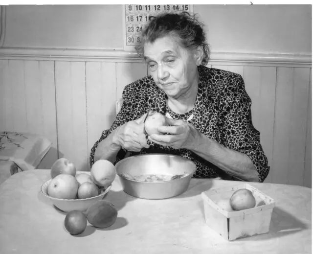 An elderly lady peels an apple at a table with a bowl of apples and a wall calendar in the background.