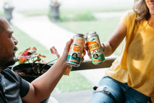 Two individuals cheer with canned beverages while sitting at a wooden table, displaying a casual, social moment.