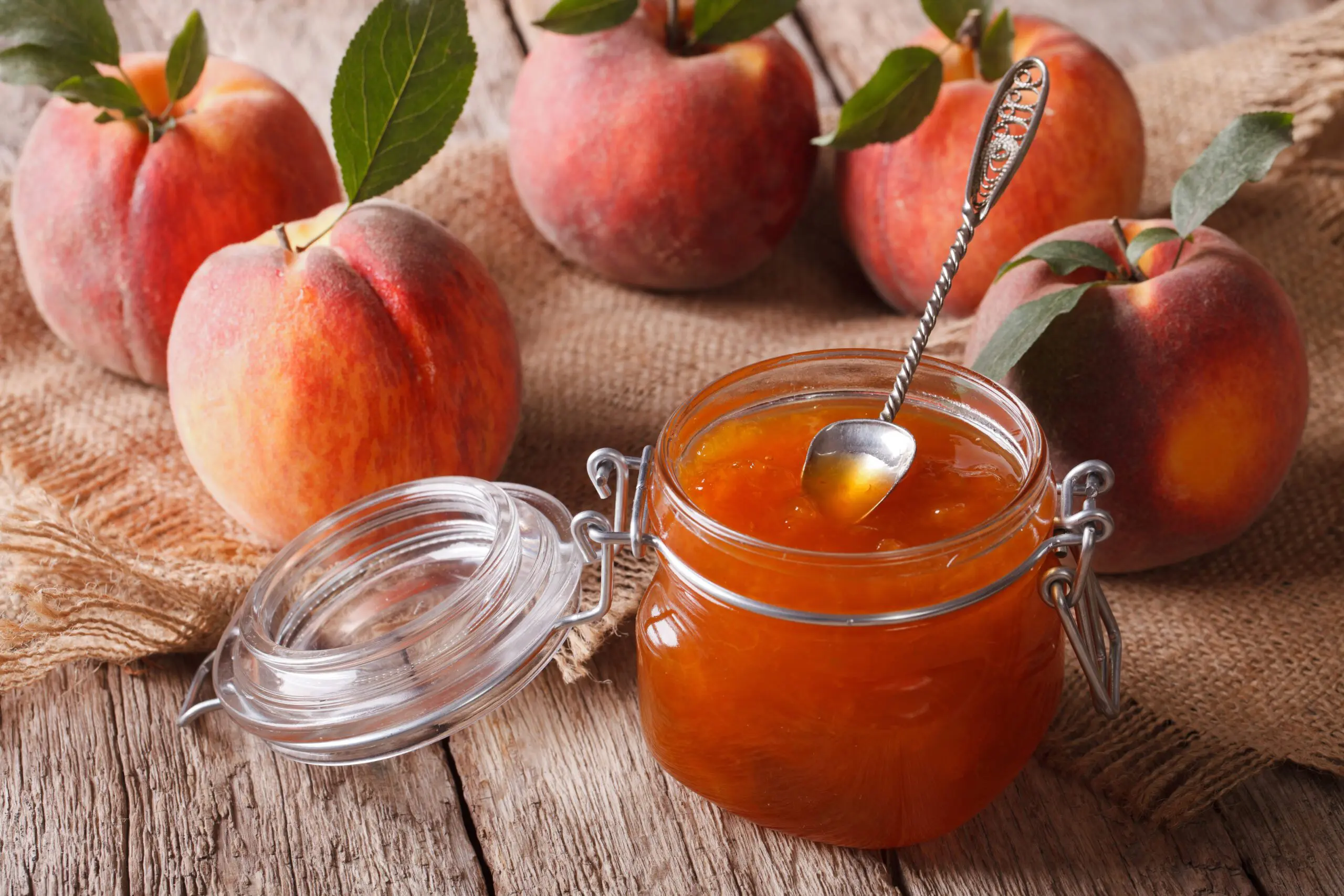 Peach Jam in an open jar with spoon and whole peaches on wooden table