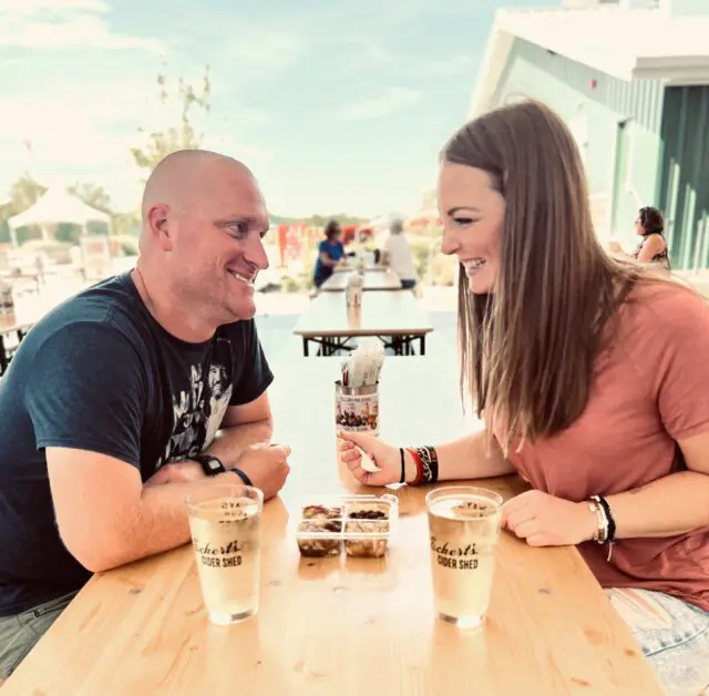 Man and woman sitting at an outdoor picnic table drinking ciders