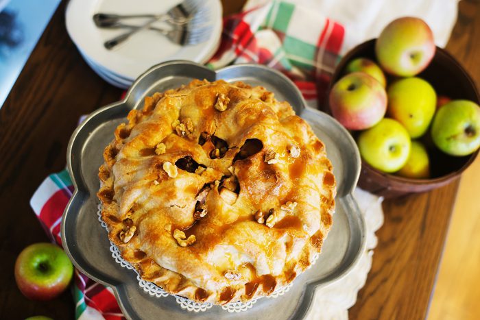 A freshly baked apple pie with a golden crust and walnuts on top, next to a bowl of multicolored apples on a wooden table with cutlery and napkin.