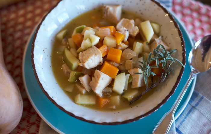 A bowl of vegetable soup with chunks of bread, garnished with herbs, next to a spoon on a patterned tablecloth.