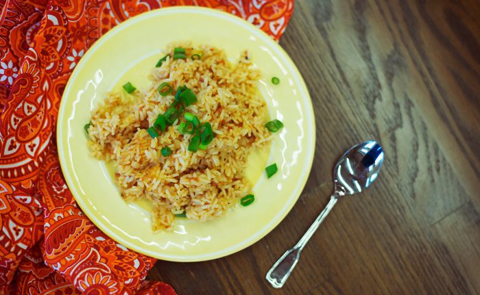 A plate of fried rice garnished with green onions next to a spoon, on a vibrant orange patterned tablecloth.