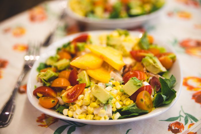 A colorful plate of salad with corn, tomatoes, mango, avocado, and greens on a floral tablecloth.