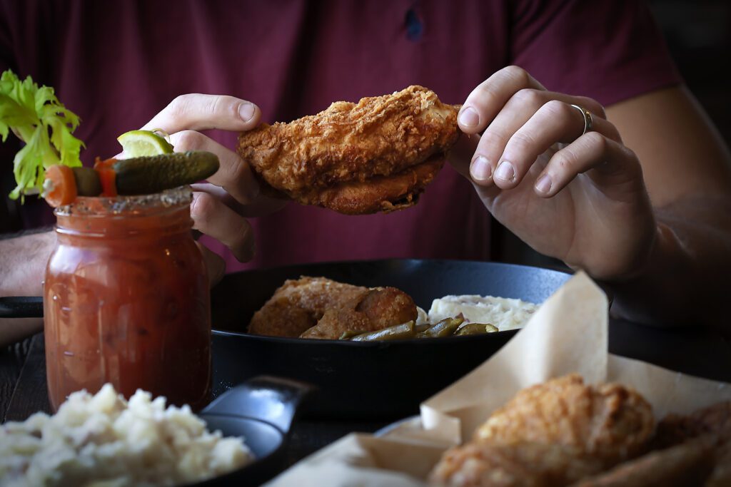 A person is holding a crispy fried chicken piece with more food and a drink on the table.