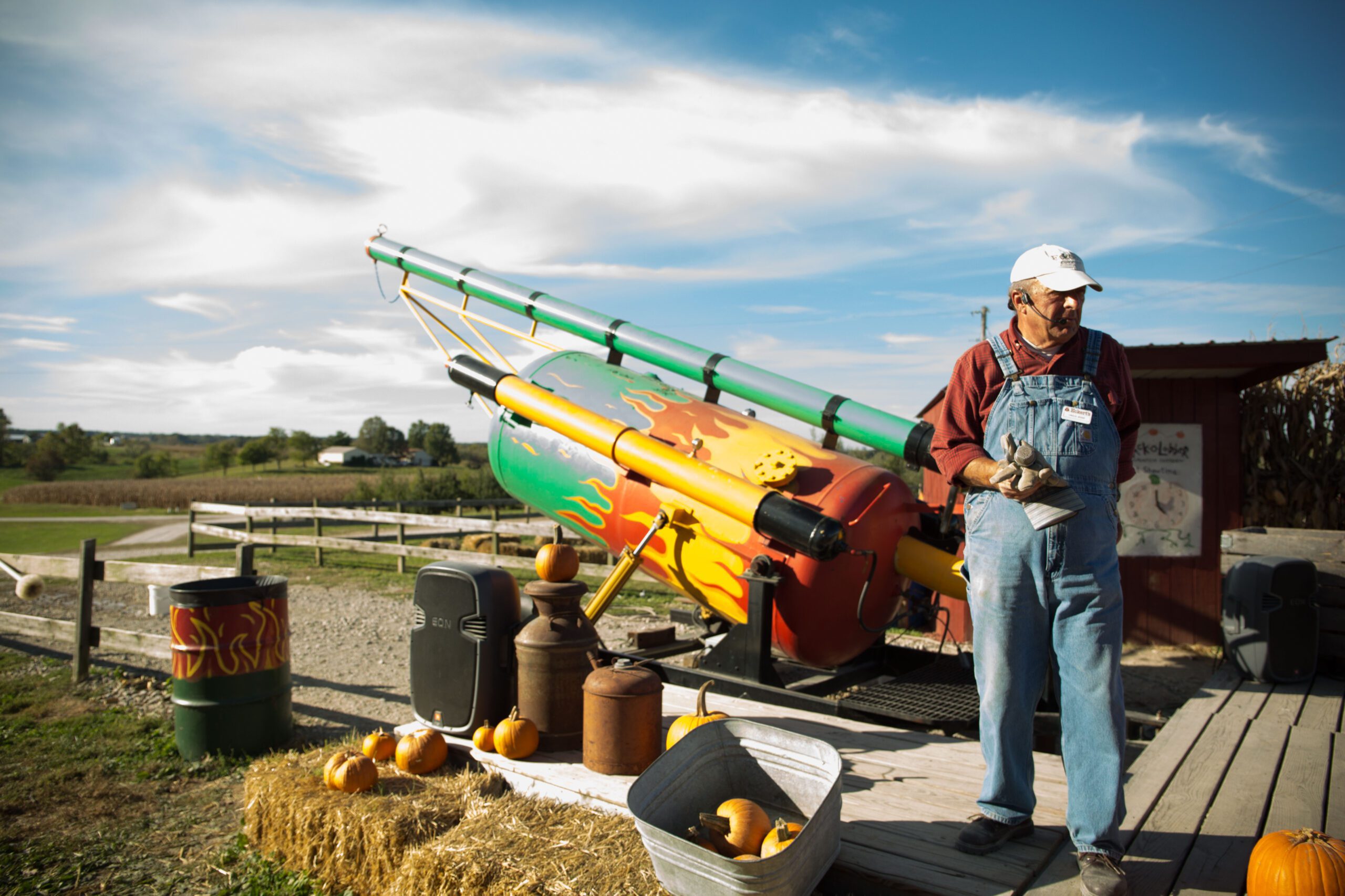 An individual stands beside a colorful pumpkin cannon on a sunny day with a clear sky.