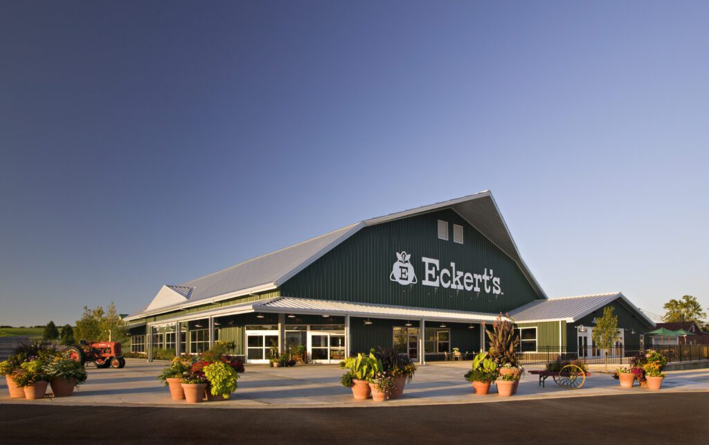 A modern building with a green roof and "Eckert's" sign under a clear blue sky flanked by potted plants.