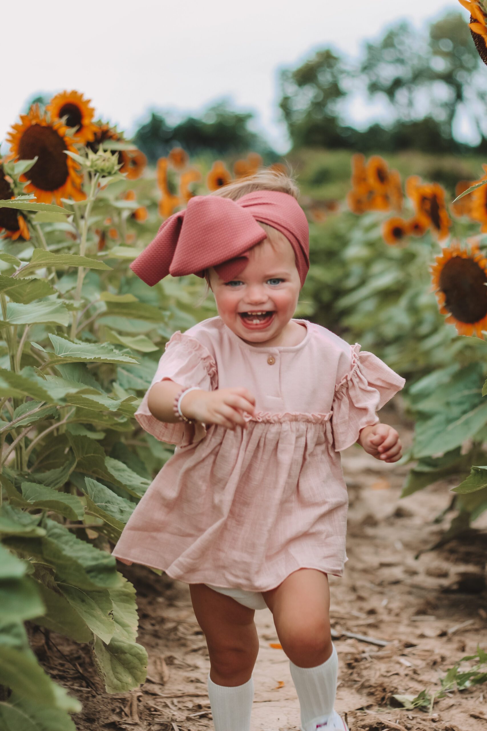 A joyous toddler in a pink dress and hat runs through a sunflower field.