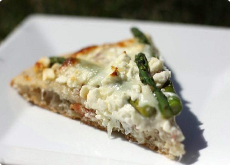 A slice of pizza with asparagus and cheese toppings on a white plate under natural light.