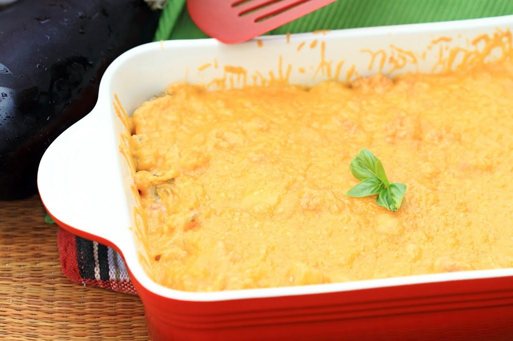 A baked cheesy dish in a red casserole served on a striped placemat with an avocado and spatula nearby.