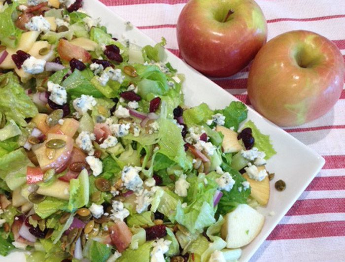 A fresh salad with apples, nuts, and blue cheese on a plate, alongside two whole apples on a striped tablecloth.