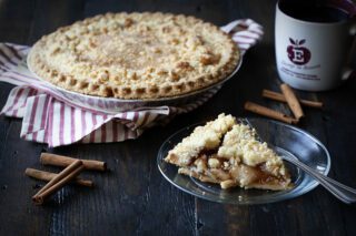 A freshly baked apple pie with a crumble topping sits next to a slice on a plate, some cinnamon sticks, and a mug.