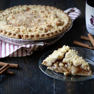 A freshly baked apple pie with a crumble topping sits next to a slice on a plate, some cinnamon sticks, and a mug.