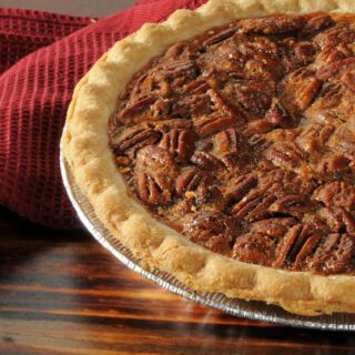 A freshly baked pecan pie sits on a wooden table next to cinnamon sticks and a red cloth.