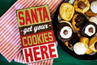 A festive sign reading "SANTA get your COOKIES HERE" alongside a plate of assorted holiday cookies on a green background.