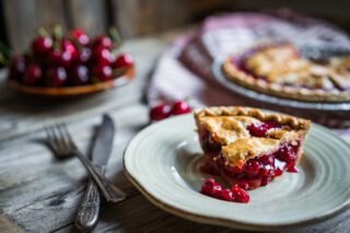 A slice of cherry pie on a plate with a full pie and fresh cherries in the background.