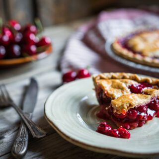 A slice of cherry pie on a plate with a full pie and fresh cherries in the background.