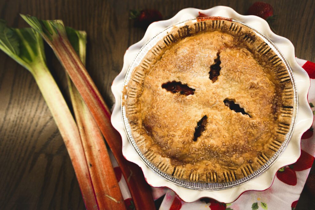 A freshly baked pie with a golden crust beside stalks of rhubarb and red petal decorations on a wooden surface.