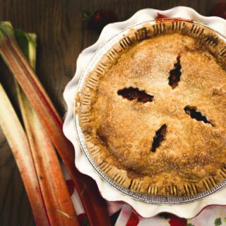 A freshly baked pie with a golden crust beside stalks of rhubarb and red petal decorations on a wooden surface.