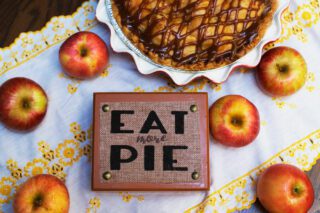 A freshly baked pie with a caramelized lattice crust is surrounded by red apples, next to a sign that says "EAT MORE PIE."