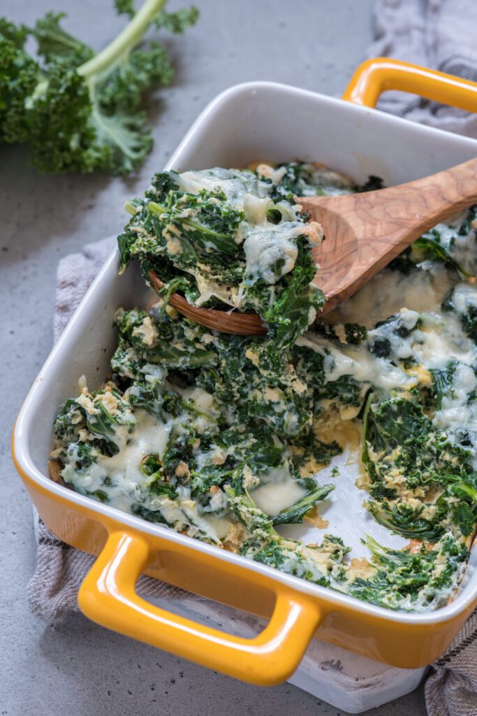 A creamy spinach and kale dish is served in a yellow-handled baking dish with a wooden spoon.