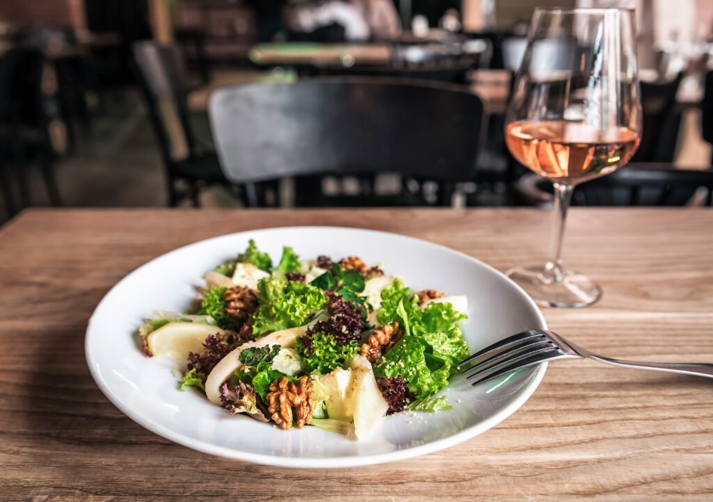 A fresh salad with greens, nuts, and cheese paired with a glass of rosé wine on a wooden restaurant table.