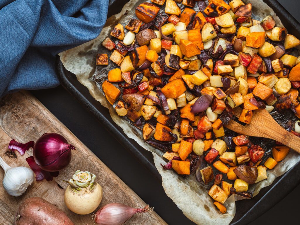 A tray of roasted root vegetables is surrounded by whole ingredients against a dark background.