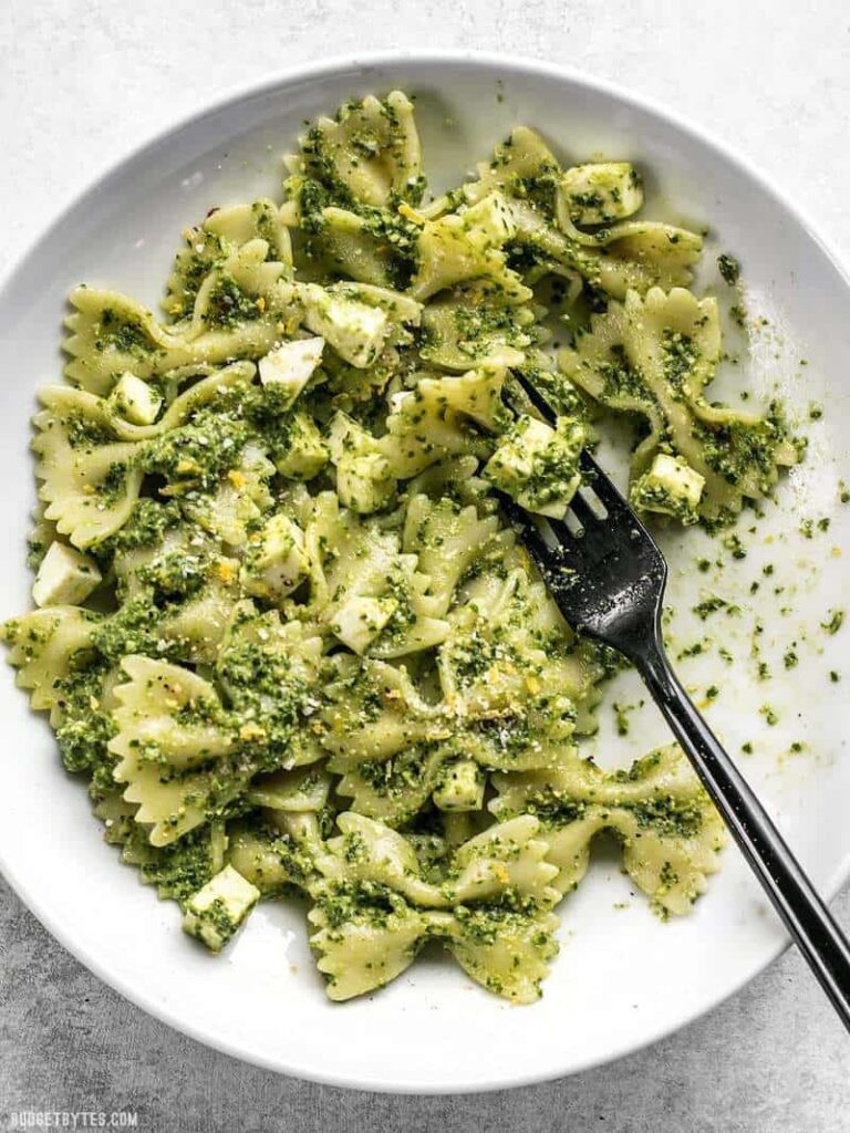 A plate of bowtie pasta with pesto sauce and cheese, garnished with herbs, accompanied by a black fork.
