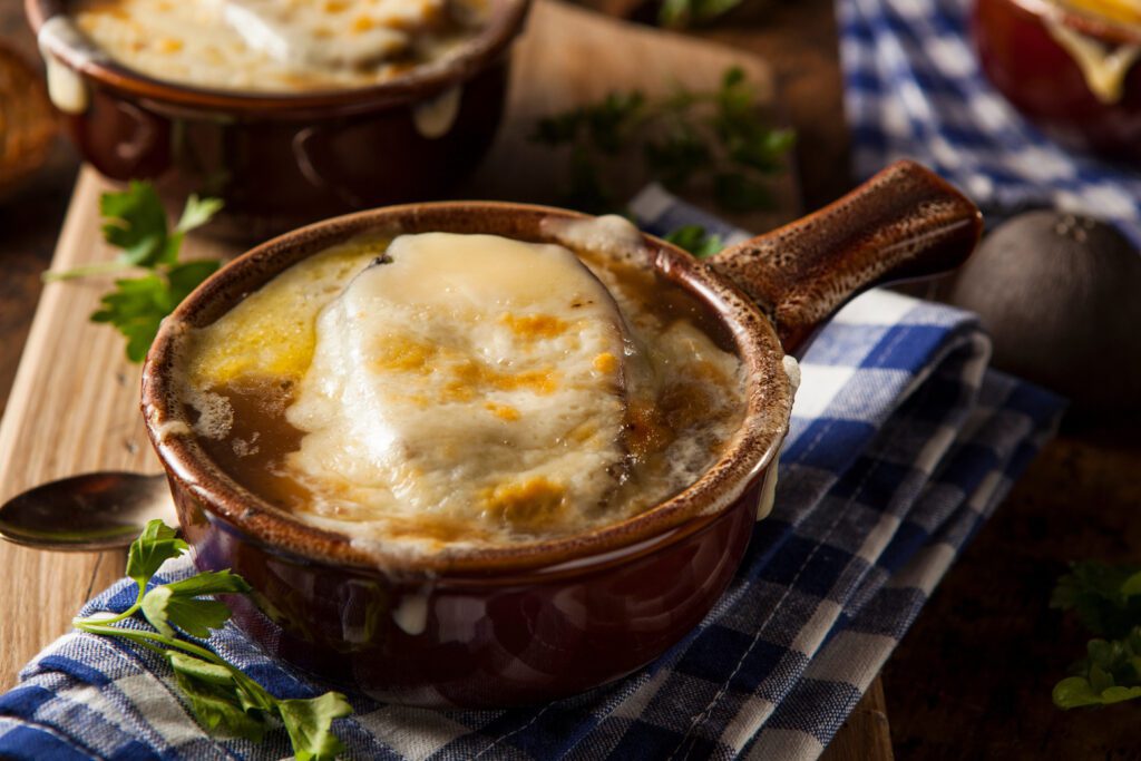 A bowl of French onion soup topped with melted cheese on a wooden table with a checkered napkin and rustic cutlery.