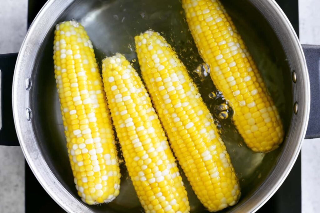 Fresh yellow corn cobs are being boiled in a silver pot of water.