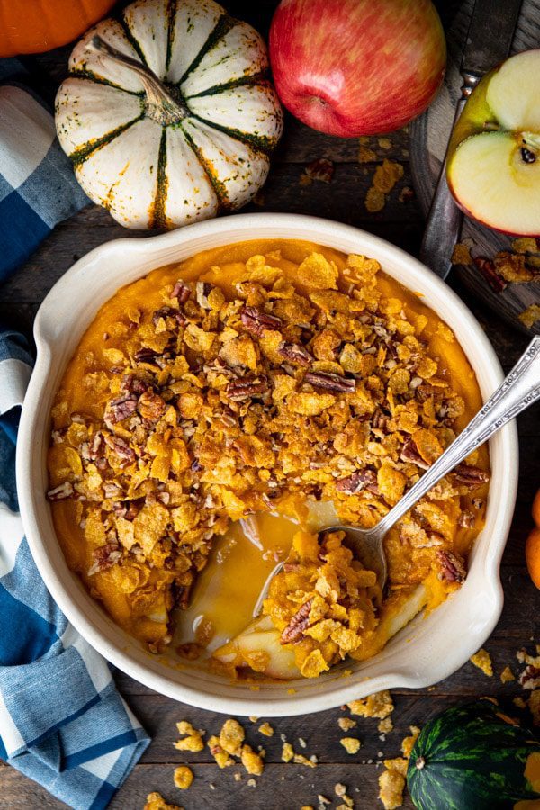 A bowl of pumpkin oatmeal topped with granola and pecans, accompanied by apples and decorative pumpkins on a wooden surface.