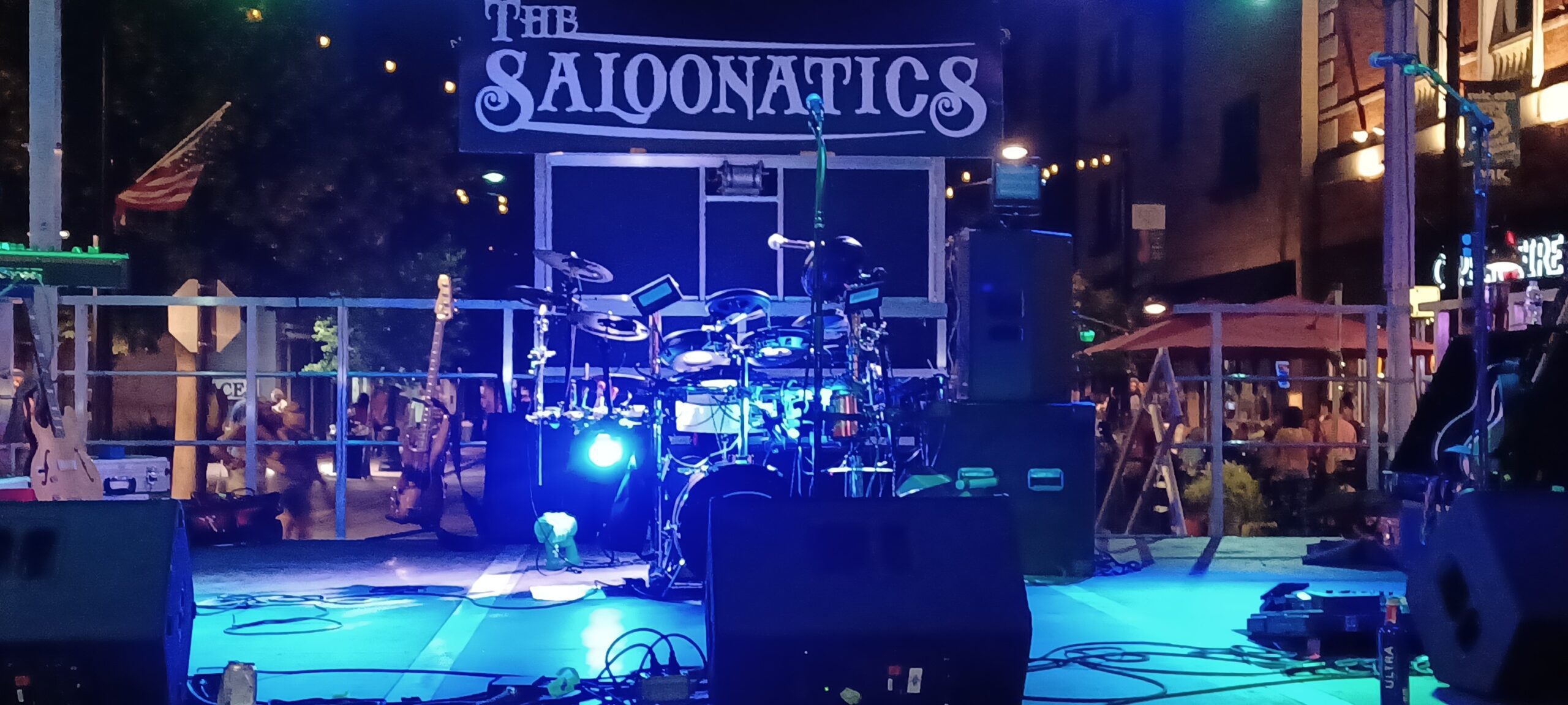 A nighttime outdoor stage setup with drums and "THE SALOONATICS" sign for an upcoming performance.