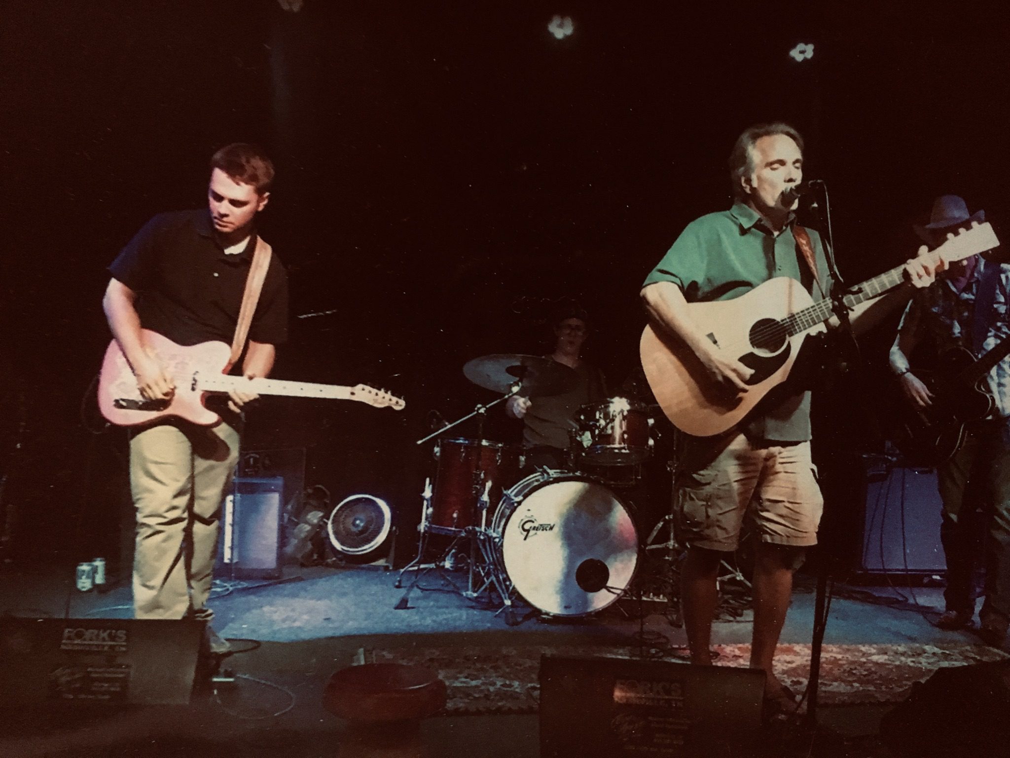 Musicians perform on stage with a drummer in the background and a guitarist and vocalist at the front.