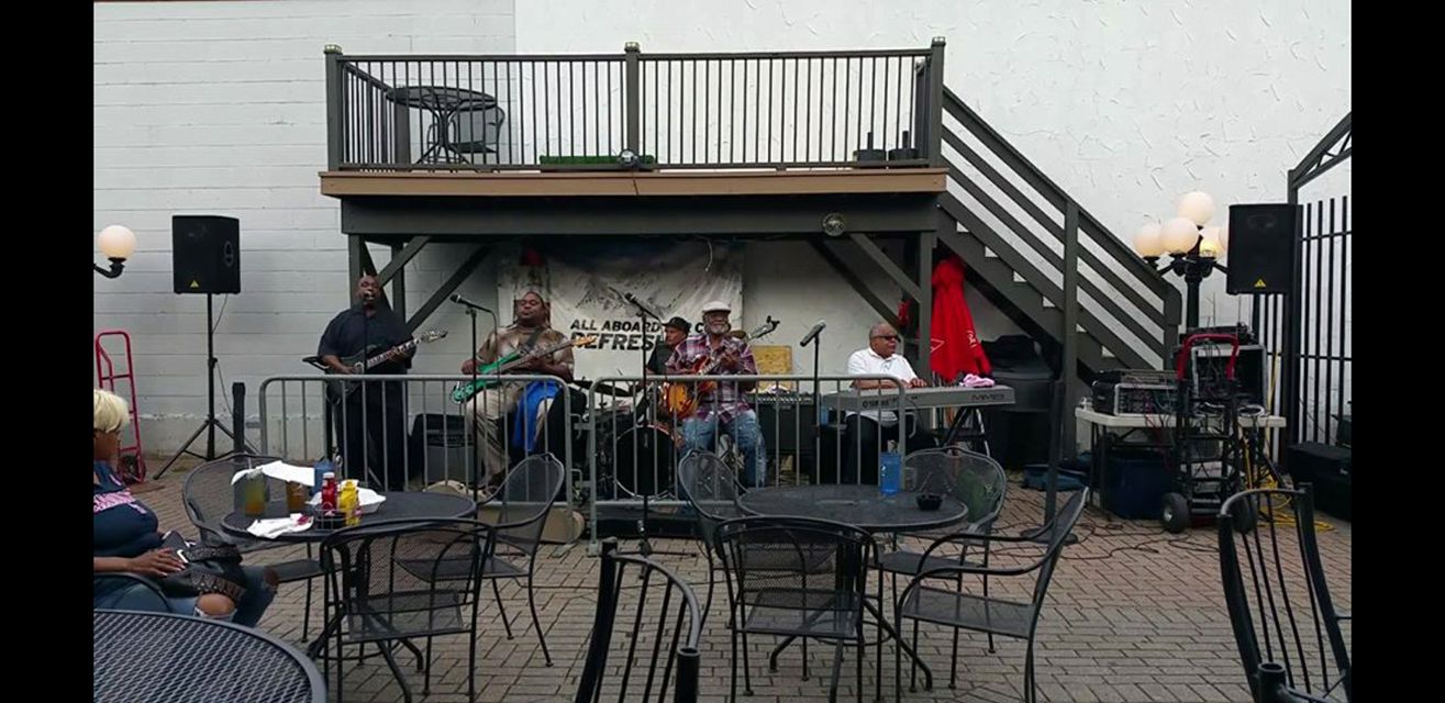 A band performs on an outdoor stage with empty chairs in the foreground and a balcony above.