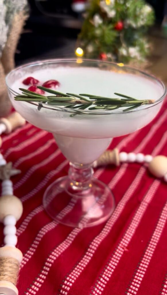 A festive cocktail garnished with rosemary and pomegranate seeds sits on a red and white striped cloth with holiday decorations in the background.